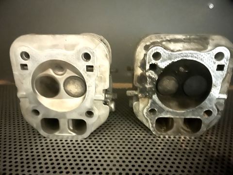 Zwei gleiche Zylinderköpfe in der Strahlkabine: links mit Walnussgranulat gereinigt und metallisch hell, rechts noch verkokt